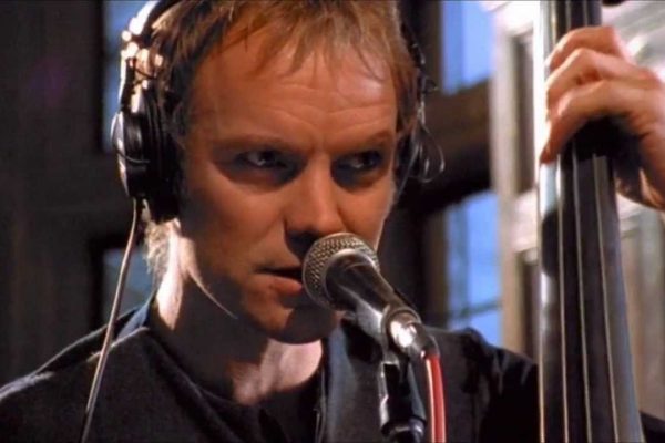 Sting, holding a bass as he sings into a microphone.